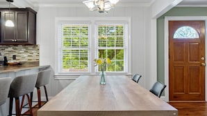 Een eethoek met een houten tafel, barstoelen, een vaas met bloemen en een raam met jaloezieën.
