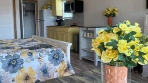A room with a bed, a wooden table, a ceiling fan, a kitchen area, and a potted plant.
