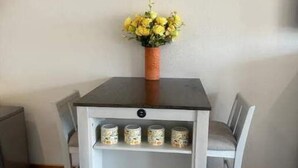 A dining table with a vase of yellow flowers, a shelf with cans, and a white chair.