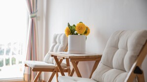 Een kamer met een houten tafel, een witte stoel en een witte bank. Op tafel staat een witte vaas met gele bloemen.