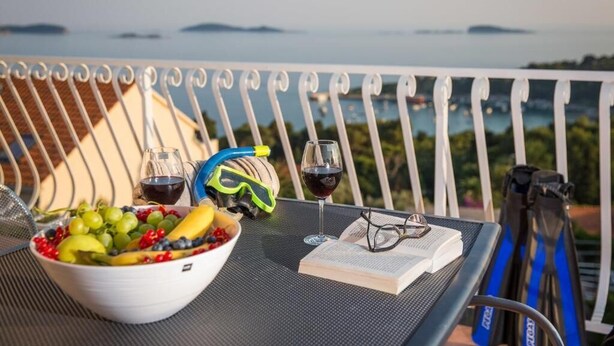 Een tafel met een fruitschaal, twee wijnglazen, een paar zwembrillen en een boek op een balkon met uitzicht op water.
