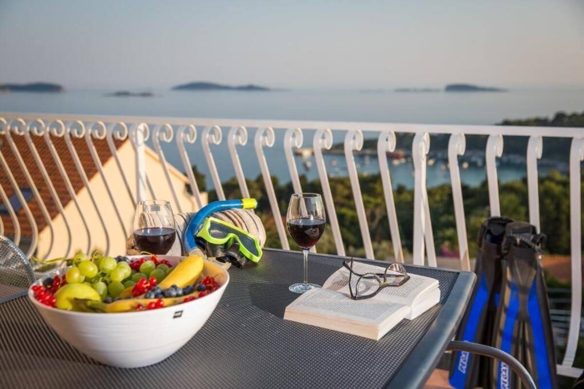 A table with a bowl of fruit, two glasses of wine, a pair of goggles, and a book on a balcony overlooking a body of water.