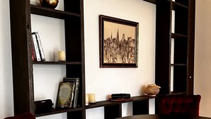 Un coin repas avec une table ronde et deux chaises à revêtement rouge, une chaise en métal noir et une table en métal noir avec un pied circulaire. Un tableau représentant un paysage urbain, encadré, est accroché au mur au-dessus de la table.