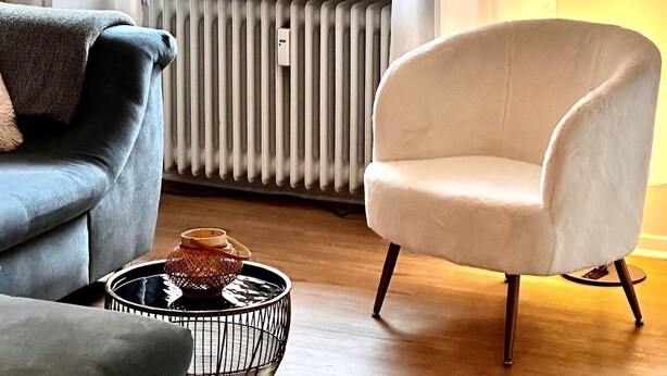 Un salon avec un canapé gris, un fauteuil blanc et une table basse en métal noir, agrémentée d’un objet décoratif.