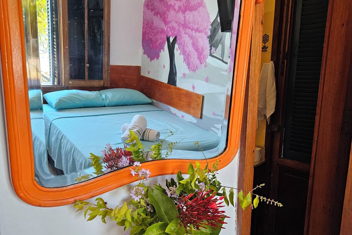 A bedroom with a bed, a mirror, a vase with flowers, and a wall with a tree mural.