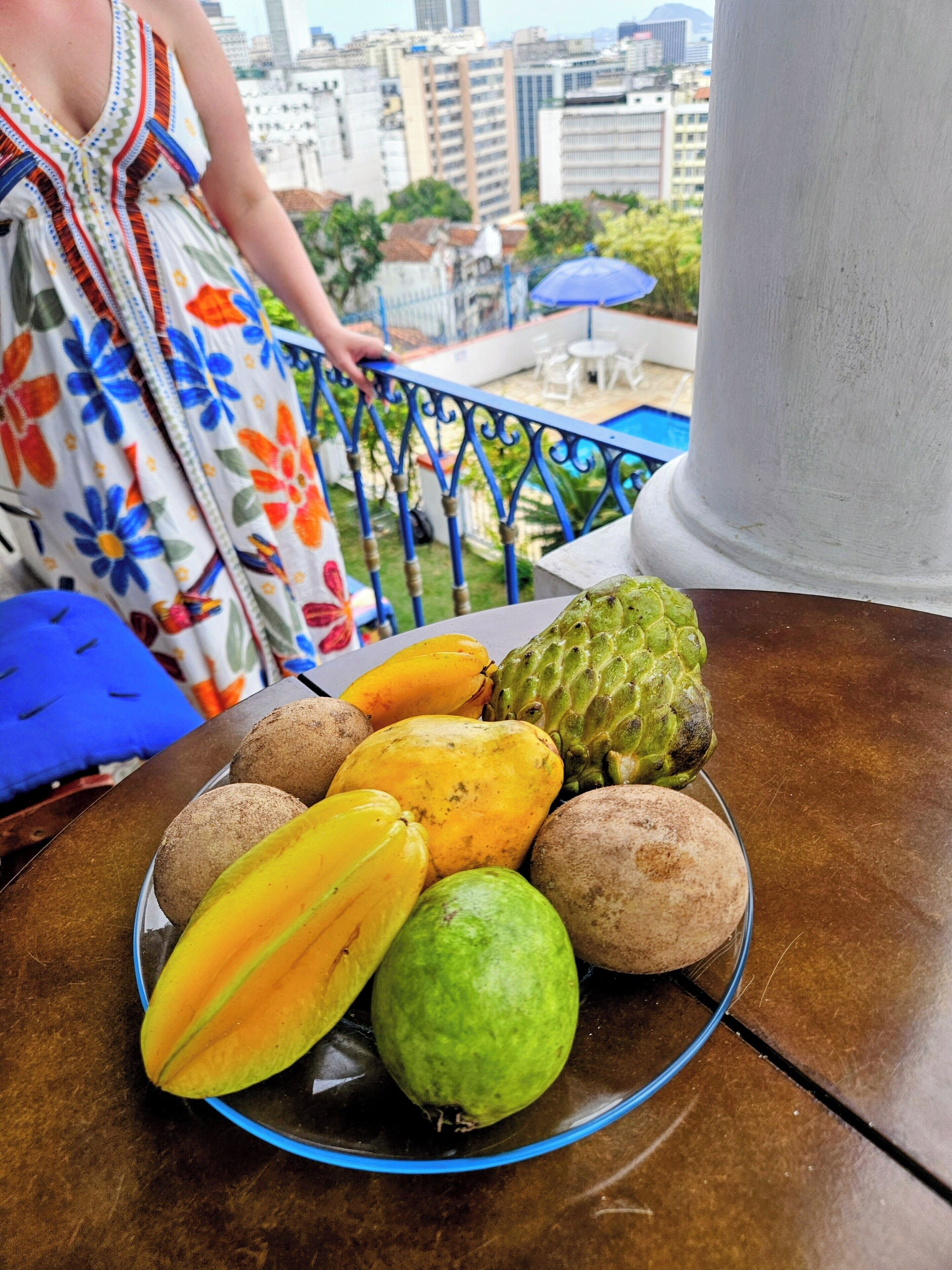 A table with a fruit bowl containing various fruits, a person in a floral dress, and a cityscape view.