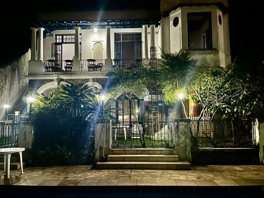 Un edificio de dos pisos con balcón, escaleras y un jardín con plantas y una mesa.