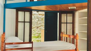 Une chambre avec des lits superposés, une tête de lit en bois, une fenêtre donnant sur un mur de pierres à l’extérieur et une rambarde bleue sur le lit supérieur.