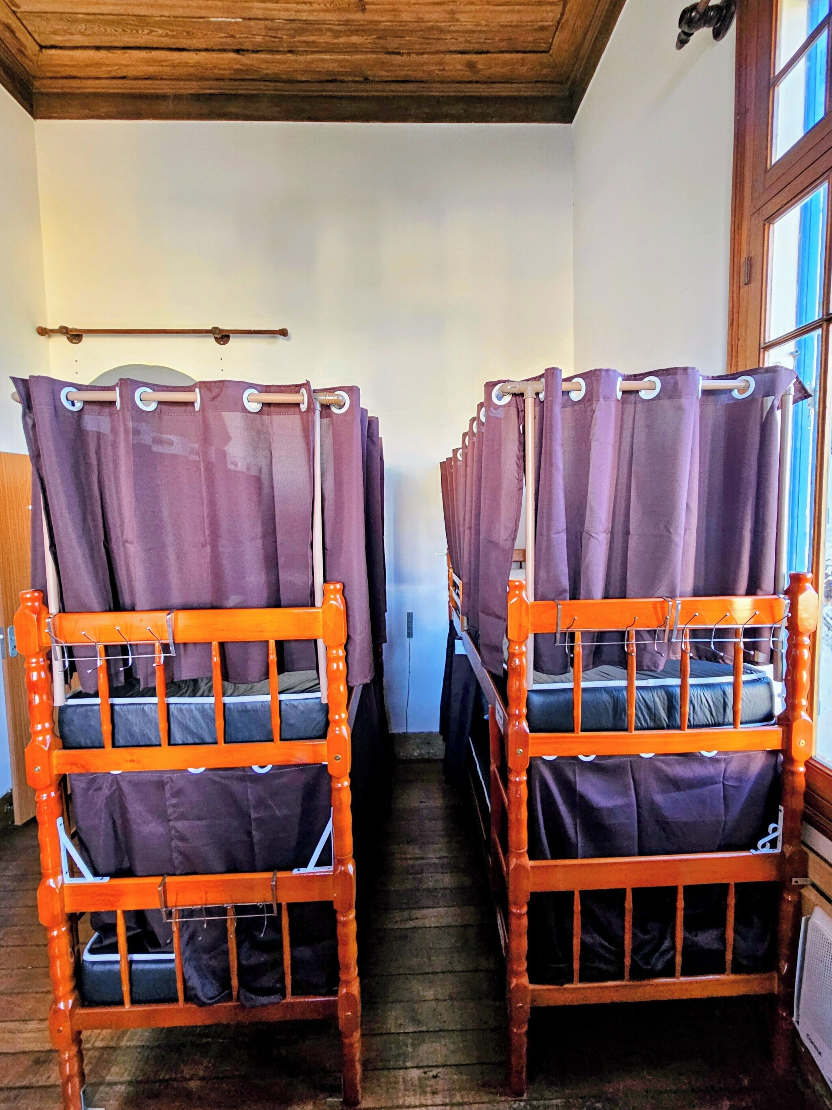 A row of wooden bunk beds with purple curtains, a wooden ceiling, and a window with blue shutters.