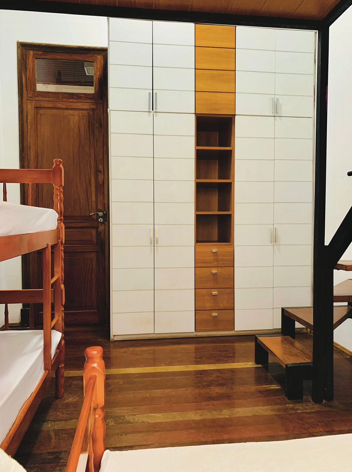Un dormitorio con una cama nido de madera, un armario con varios compartimentos y un pequeño taburete de madera.