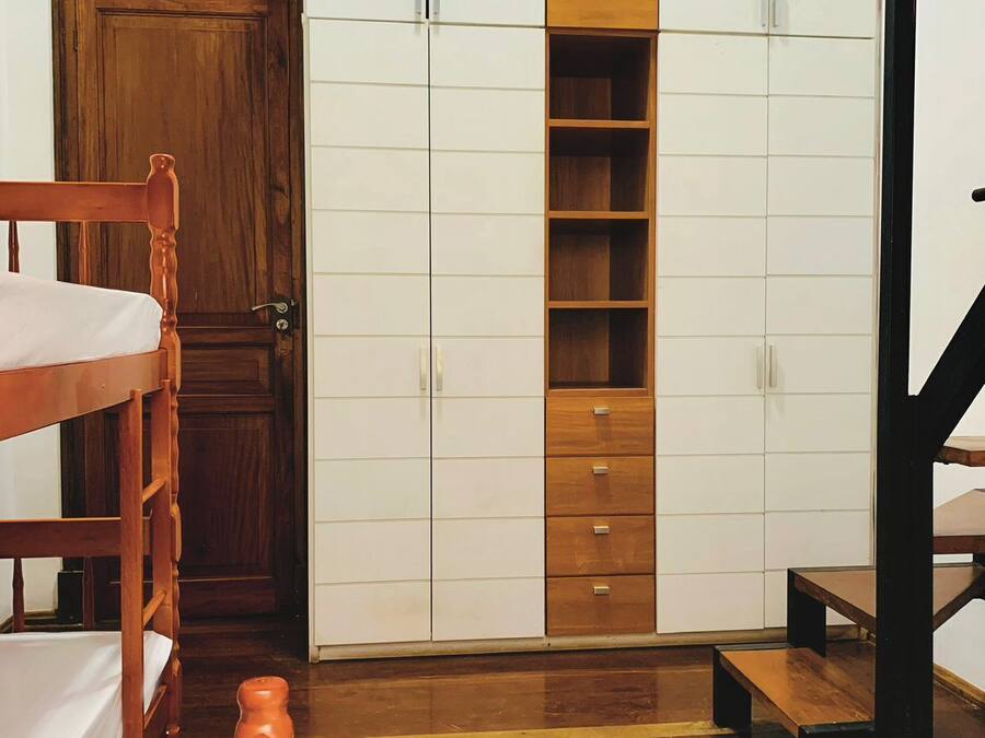 Un dormitorio con una cama nido de madera, un armario con varios compartimentos y un pequeño taburete de madera.