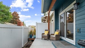 A patio with two wicker chairs and a small table, a white fence, and a blue building with a sliding glass door. - New 2 BR Townhouse in the Heart of Wenatchee. Great for Families, pet friendly. (Wenatchee)