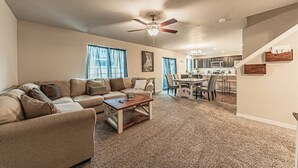 A spacious living room with a large sofa, a coffee table, and a ceiling fan. A kitchen is visible in the background with a dining area. - •Lamine Elegance Drive• FreeWine, GameRoom & HotTub (Colorado Springs)