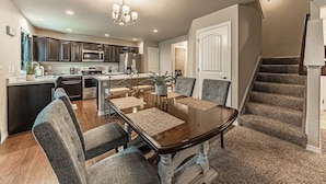 A modern kitchen with dark cabinets, stainless steel appliances, and a dining area with a wooden table and grey upholstered chairs. - •Lamine Elegance Drive• FreeWine, GameRoom & HotTub (Colorado Springs)