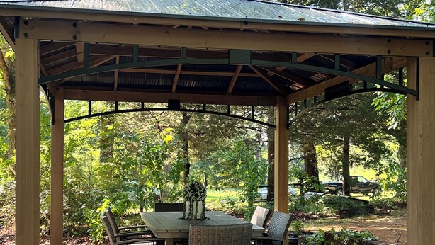 Un patio abrité, doté d’une table et de chaises, entouré d’arbres et bordé d’un chemin de gravier.
