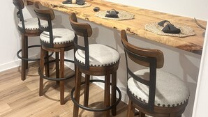 Un espace bar moderne doté d’un long comptoir en bois, de quatre tabourets de bar et d’un téléviseur à écran plat fixé au mur.