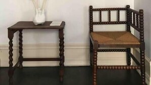 Una stanza con una sedia in legno scuro, un tavolino con un vaso bianco contenente fiori secchi e uno specchio rotondo appeso al muro.