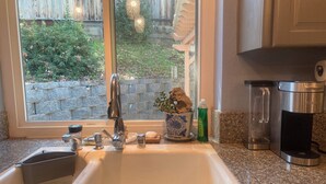 A kitchen with a granite countertop, a stainless steel sink, a coffee maker, and a window with a view of a stone wall and greenery. - Cozy 5BR Ranch Home with Hot Tub in Quiet Folsom Neighborhood (Folsom)