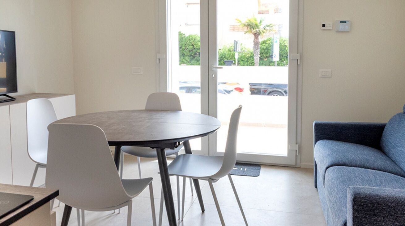 A modern living room with a round table and chairs, a sofa, a TV, and a sliding glass door leading to a parking area.