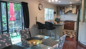 A modern kitchen with a gas stove, a microwave, and a stainless steel range hood. There is a dining area with a glass table and chairs, and a large window with blinds. - Luxury Large 2 Storied Villa Walk to Shopping Mall Howick (Auckland)