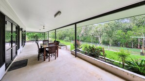 A covered patio with a table and chairs, large windows, and a view of a garden and trees. - Riverside Homestead (Darwin River)