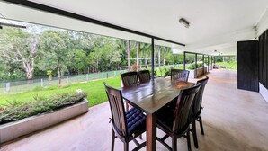 A modern outdoor dining area with a wooden table and chairs, surrounded by glass walls offering a view of a lush garden and a tennis court. - Riverside Homestead (Darwin River)