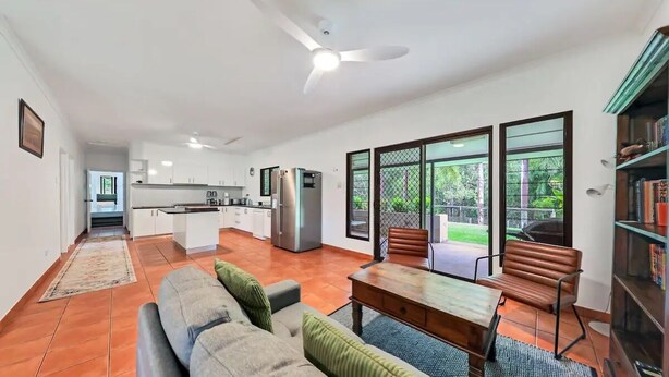 A modern living room with a sofa, a coffee table, and a bookshelf. There is a kitchen area with a refrigerator and a dining table.
