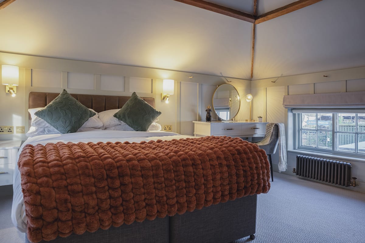 Ein Schlafzimmer mit einem großen Bett, zwei Kissen, einem Nachttisch, einer Kommode, einem Spiegel und einem Fenster.