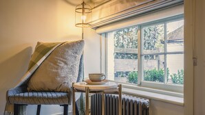 A cozy room with a cushioned chair, a small round table with a cup, and a view through a window. - The Crown Inn at Burnham Beeches (Slough)