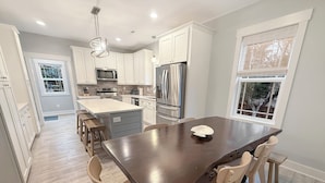 A modern kitchen with white cabinets, stainless steel appliances, a central island with stools, and large windows.
