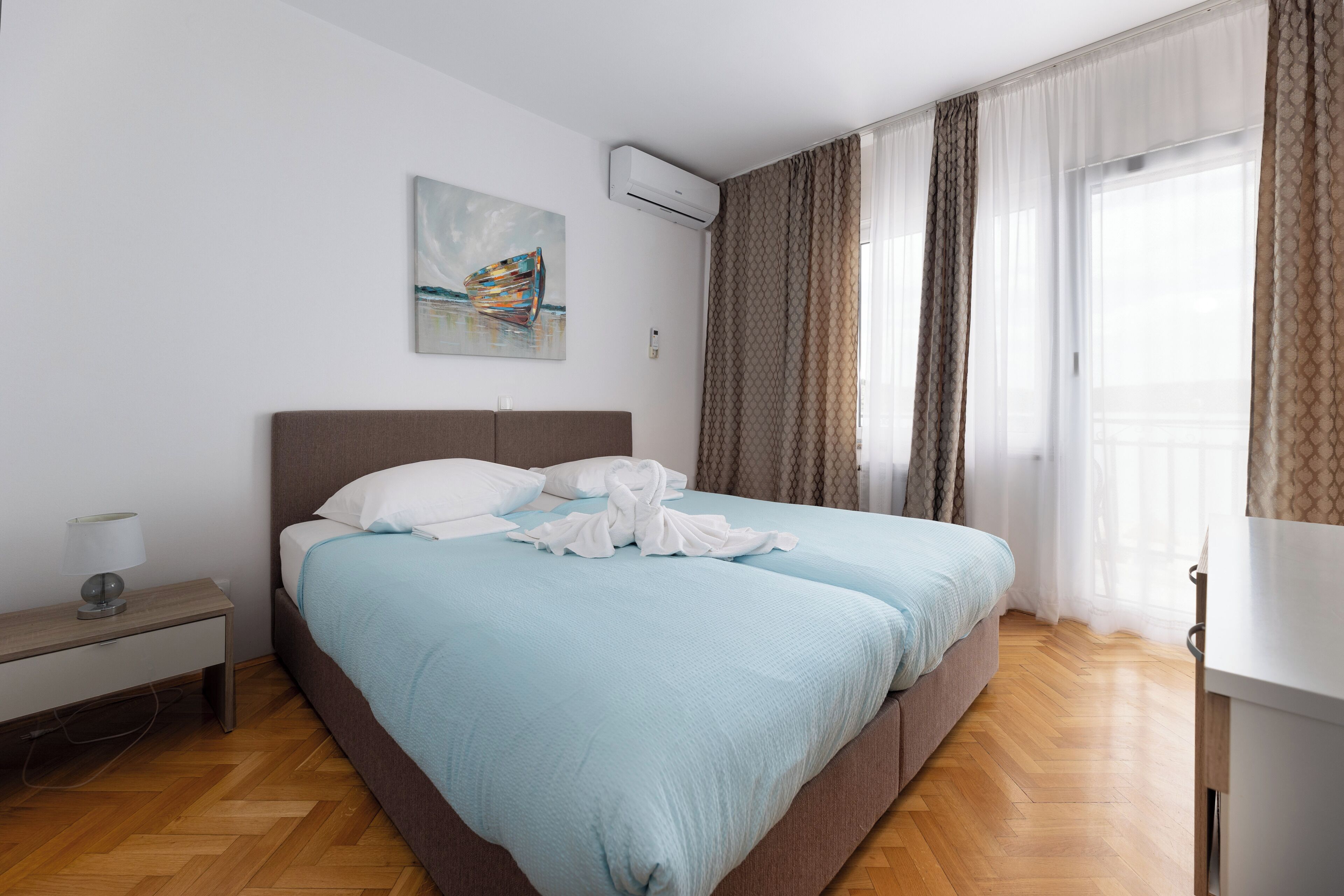 A neatly made bed with a blue bedspread and white pillows, a bedside table with a lamp, a painting on the wall, and a window with curtains.