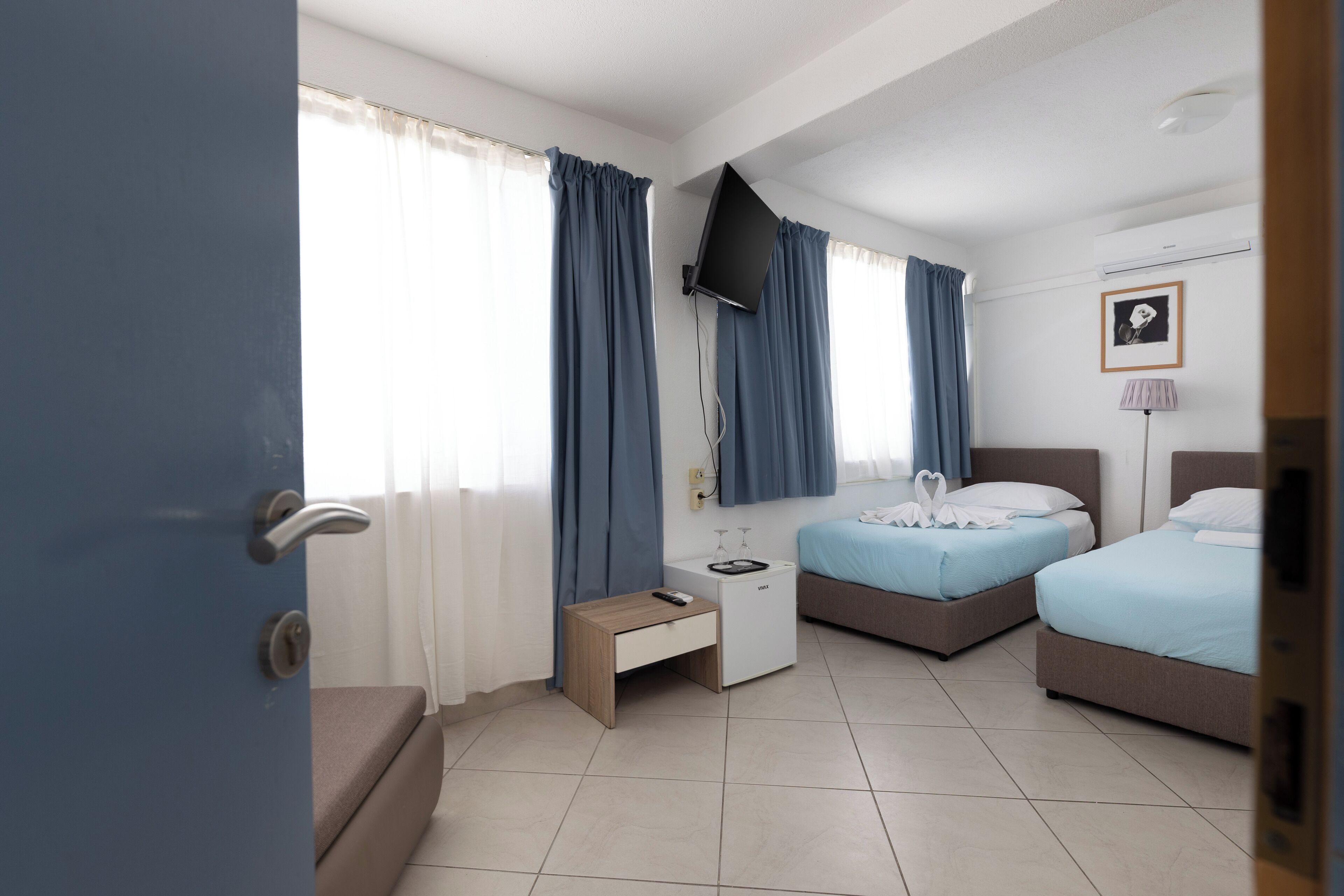 Quarto de hotel com duas camas, uma mesinha de cabeceira, TV fixada na parede e uma janela com cortinas.