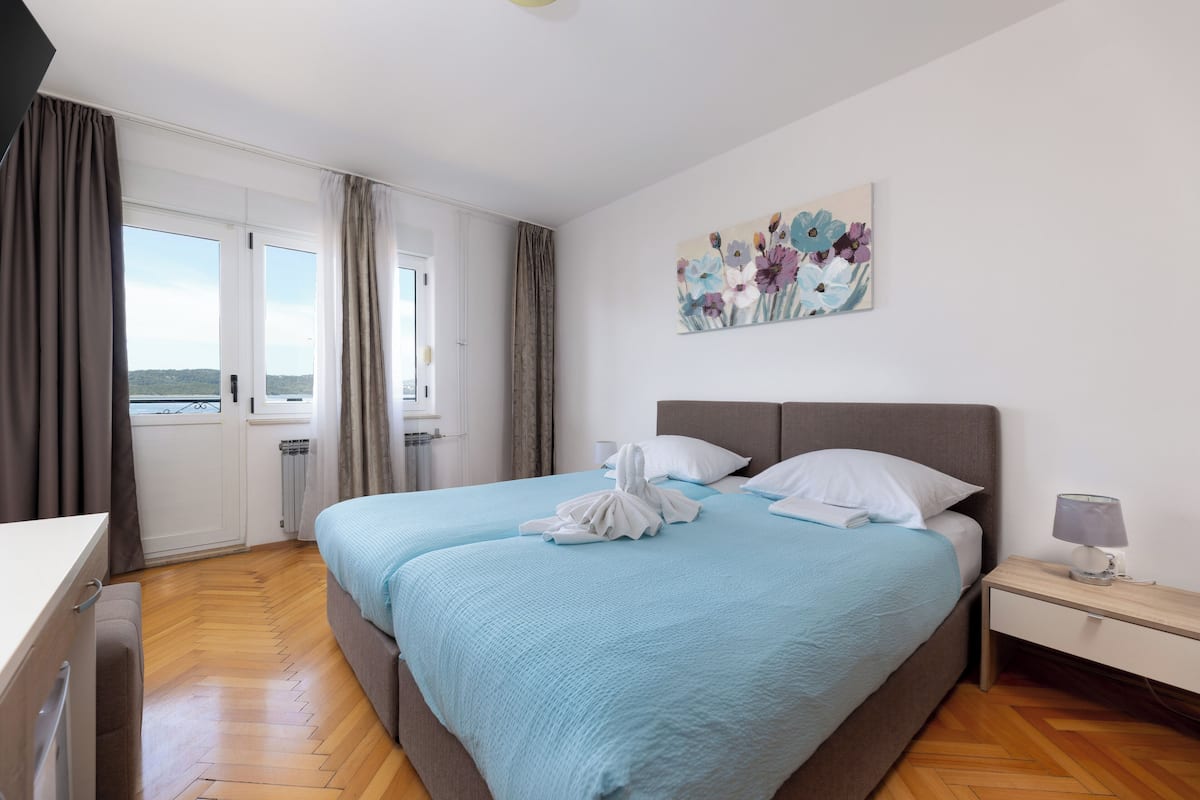 A hotel room with a bed, a nightstand, a window with curtains, and a painting on the wall.
