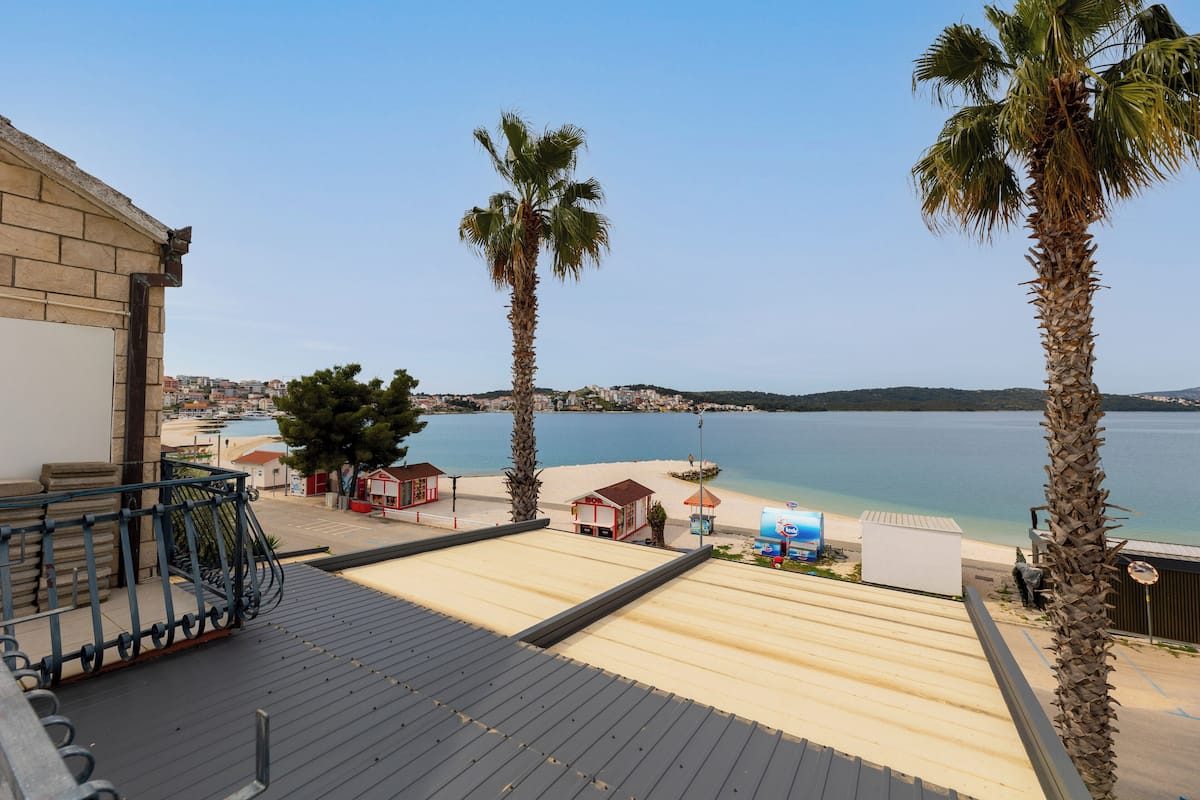 A beachside view from a balcony overlooking a sandy beach, palm trees, and a calm sea.
