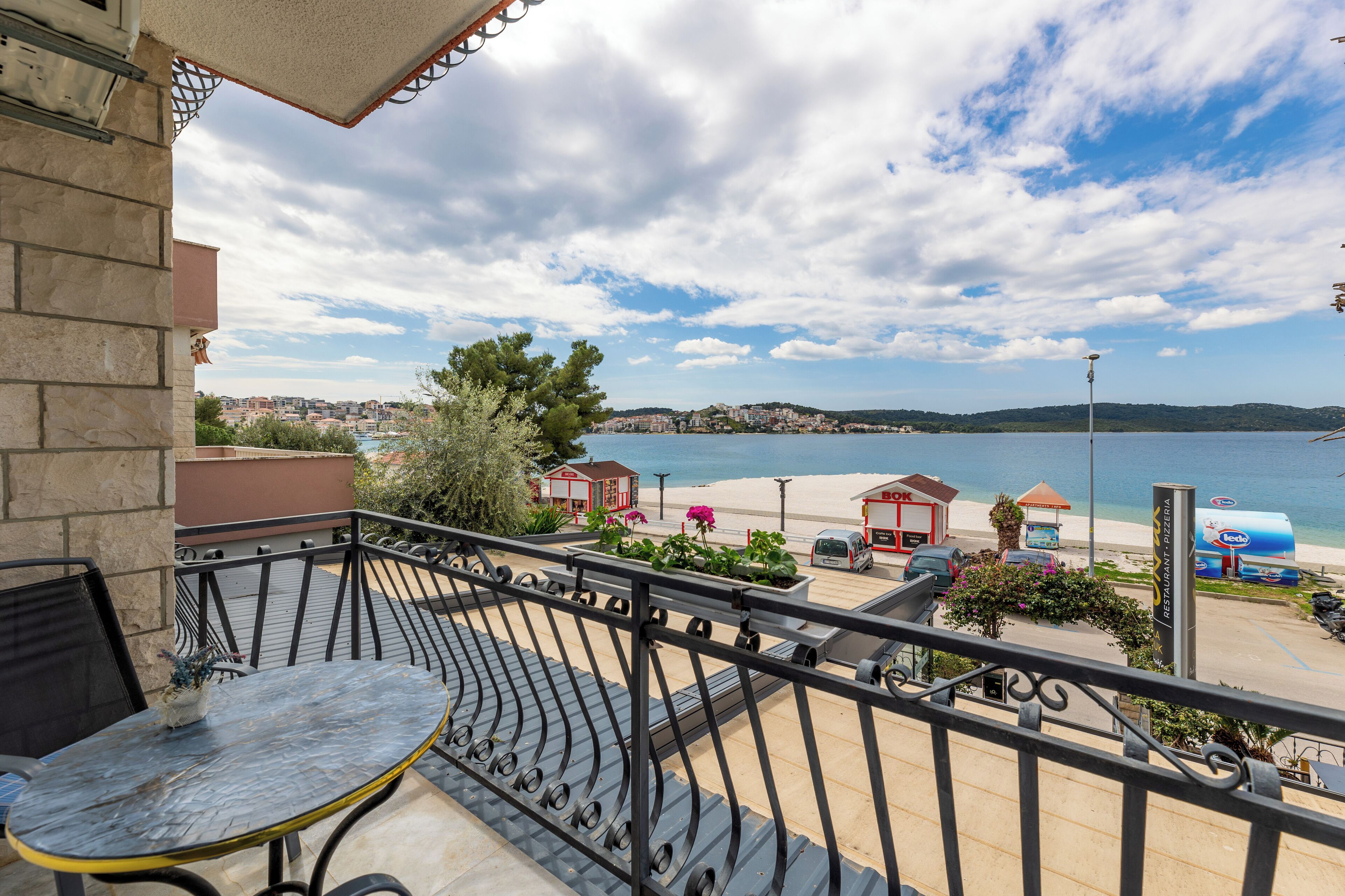 A balcony with a table and chairs, overlooking a beach with buildings and a clear sky.