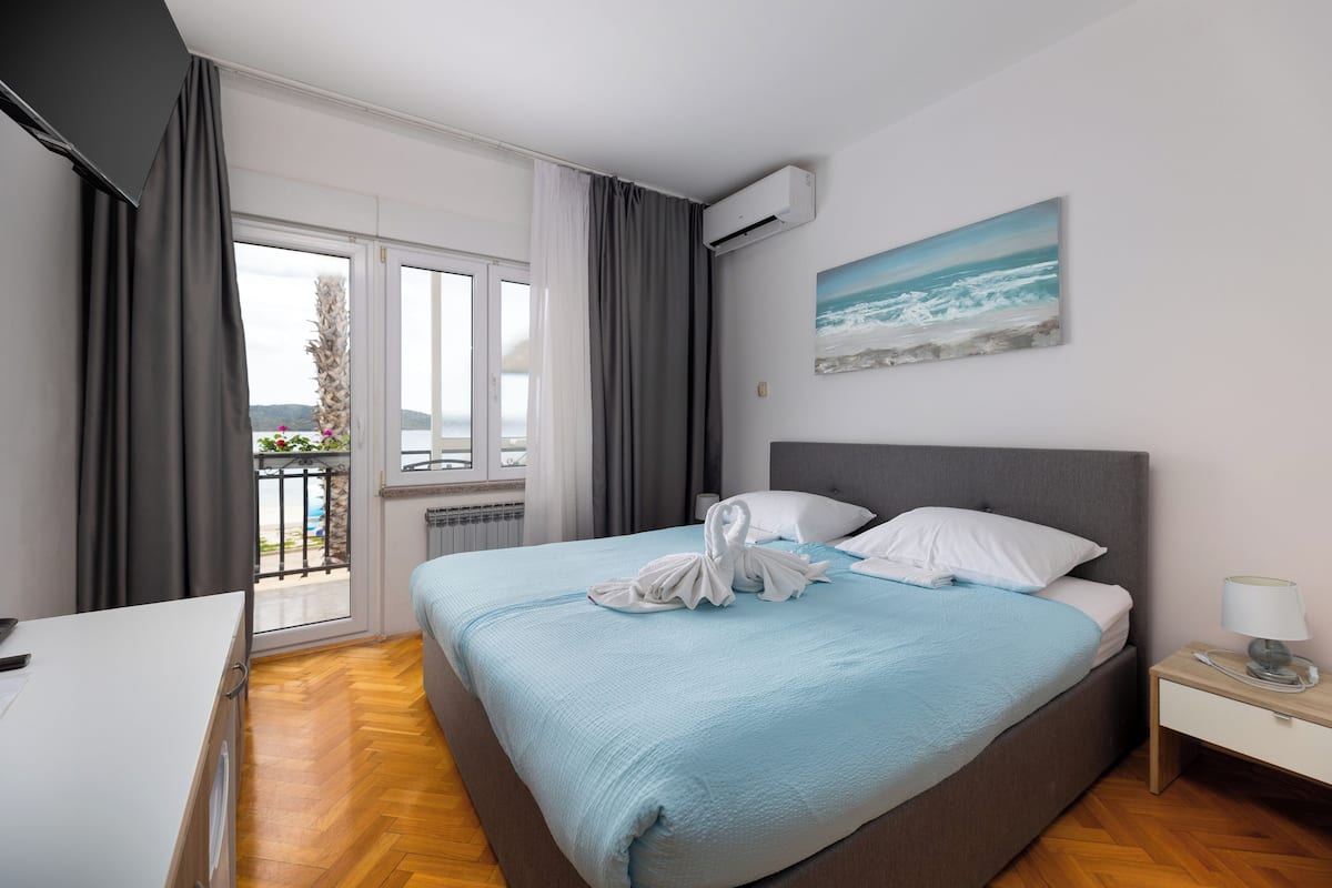 A hotel room with a bed, a nightstand, a window with curtains, and a painting of the sea.