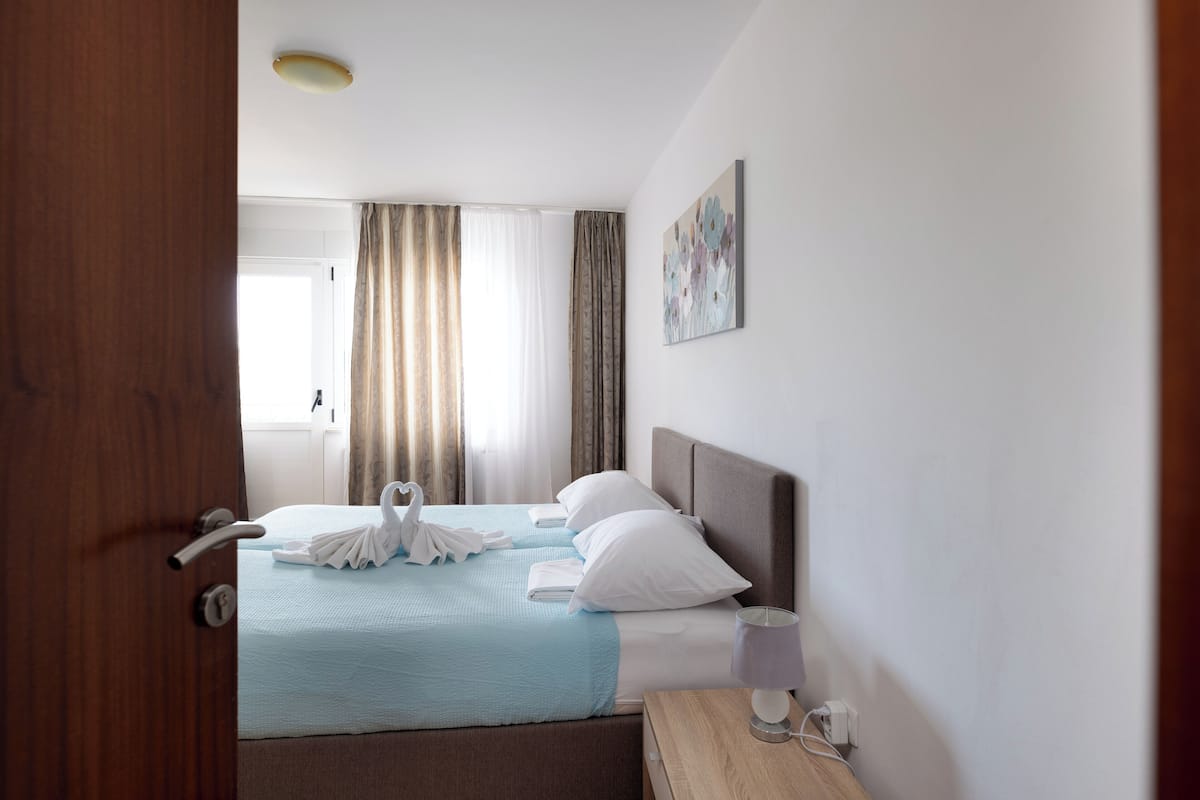 A hotel room with a bed, a bedside table, a lamp, a painting on the wall, and a window with curtains.