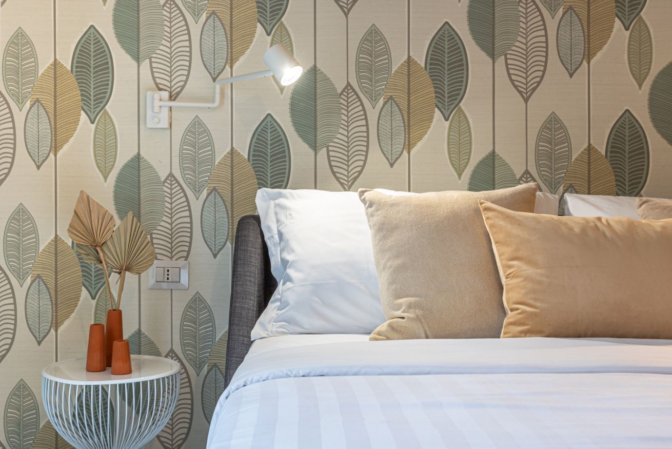 Une chambre à coucher avec un lit, des oreillers, une table de chevet avec une lampe et un mur orné d’un motif de feuilles.