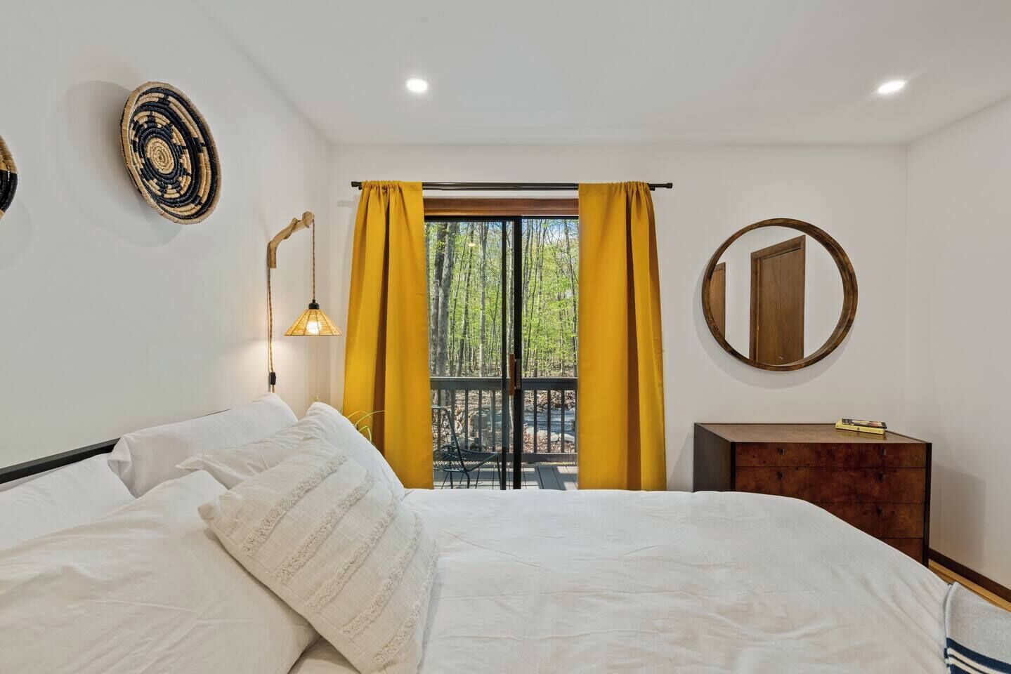 A bedroom with a bed, a nightstand, a mirror, and a window with yellow curtains.