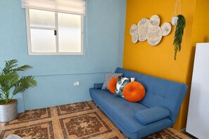 A room with a blue sofa, patterned floor tiles, a window with blinds, a wall-mounted decorative plate, and a hanging plant.