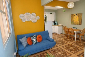 Uma sala de estar com um sofá azul, uma área de jantar com cadeiras de madeira, uma cozinha com armários brancos e uma parede amarela.