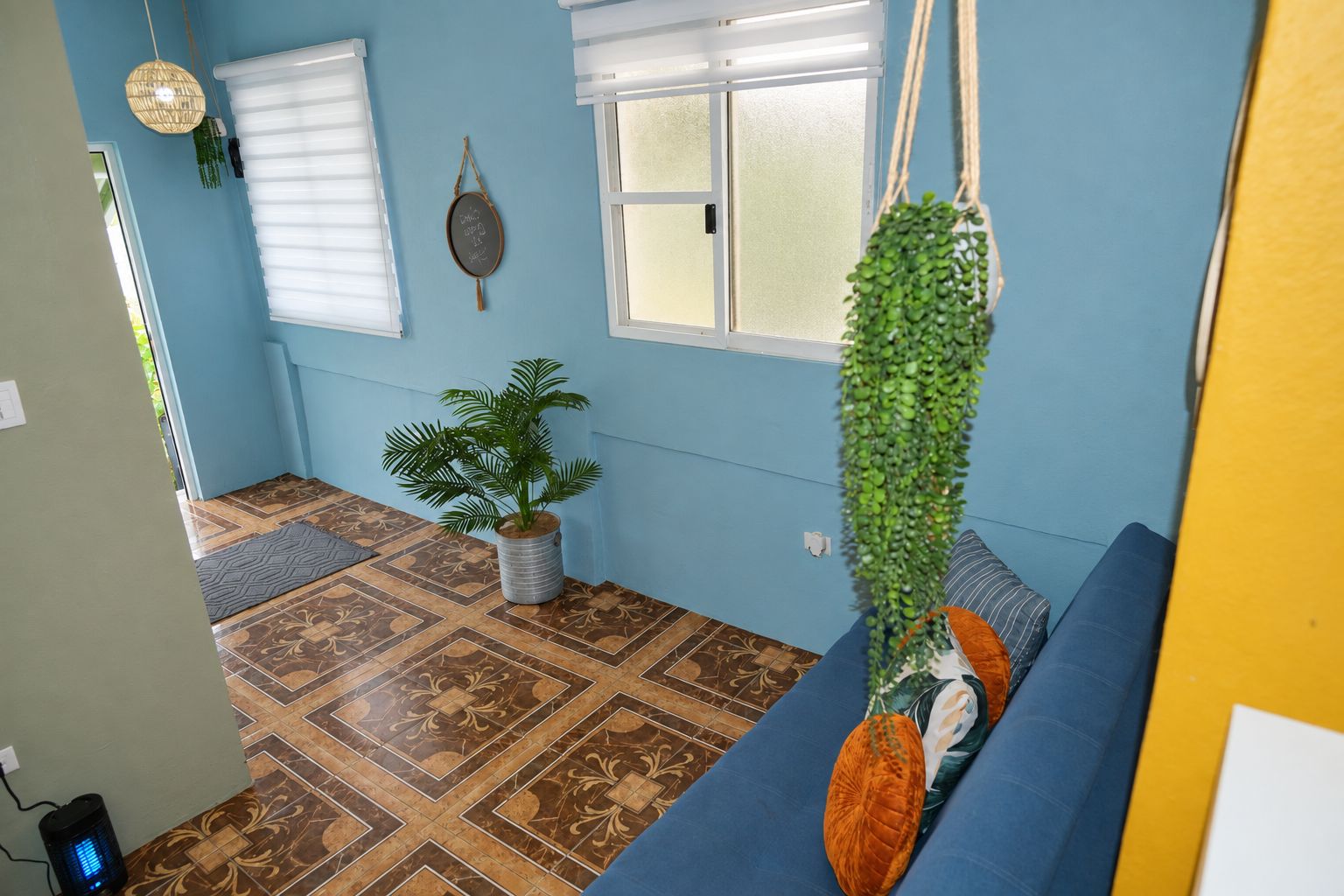 Ein Zimmer mit einem gemusterten Boden, einem blauen Sofa, einer hängenden Pflanze und einem wandmontierten Spiegel.
