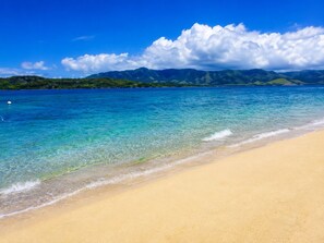 A beach with clear turquoise water, a sandy shore, and a mountain range in the background.