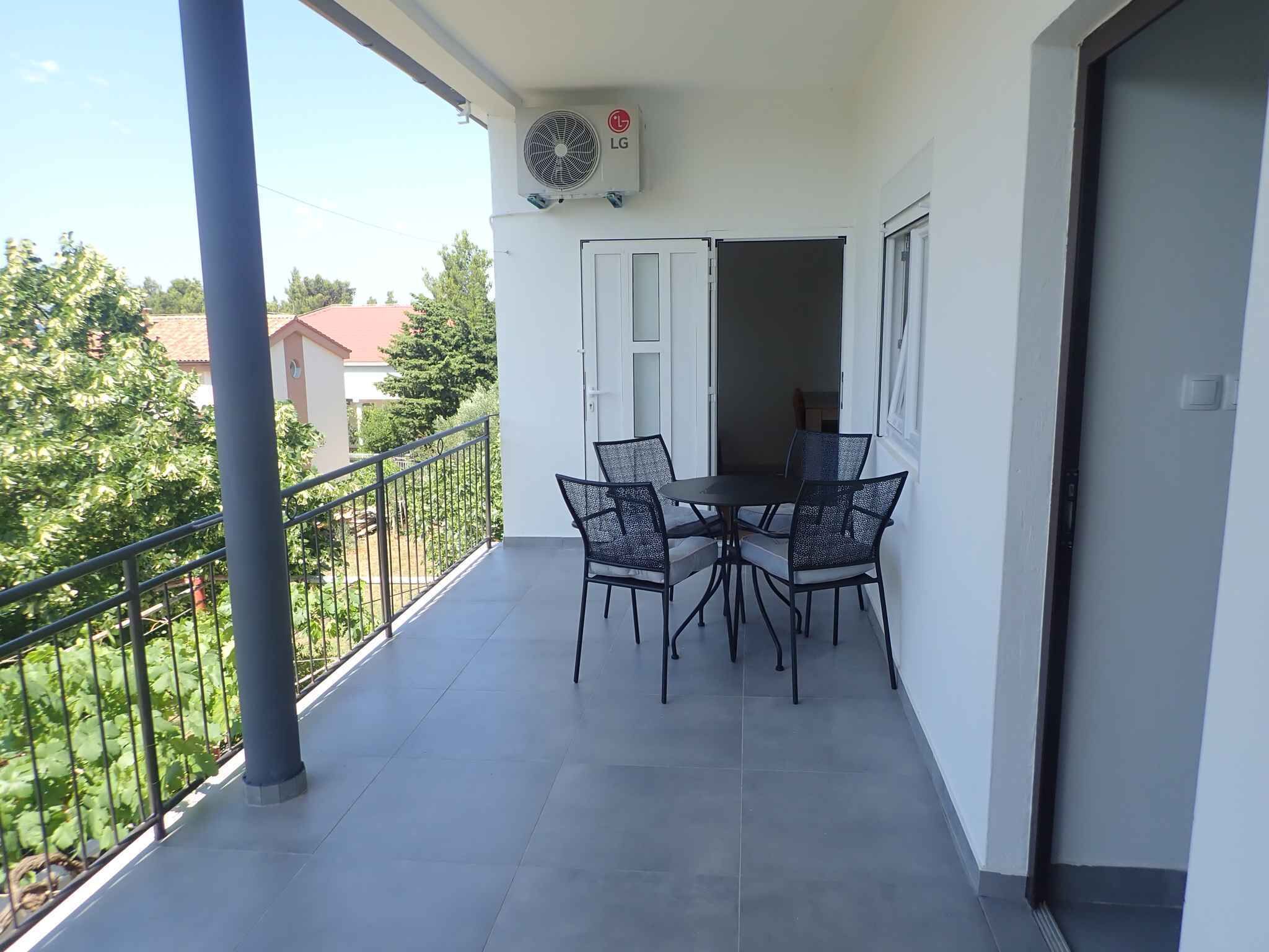 En balkon med et bord og stole, en klimaanlæg og udsigt til vegetation og bygninger.