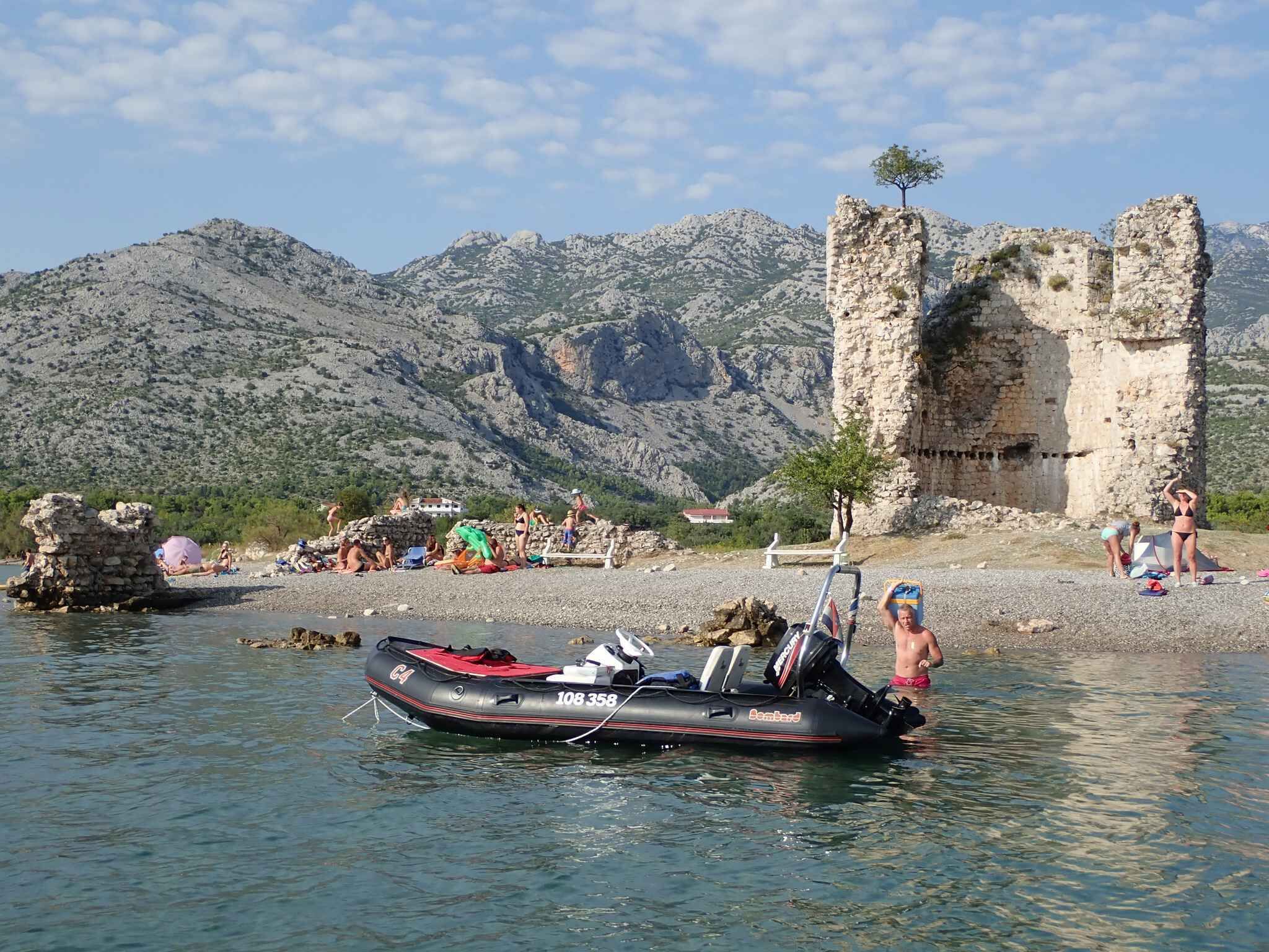 A boat on calm water near a rocky shore with people sunbathing and a historical stone structure in the background.