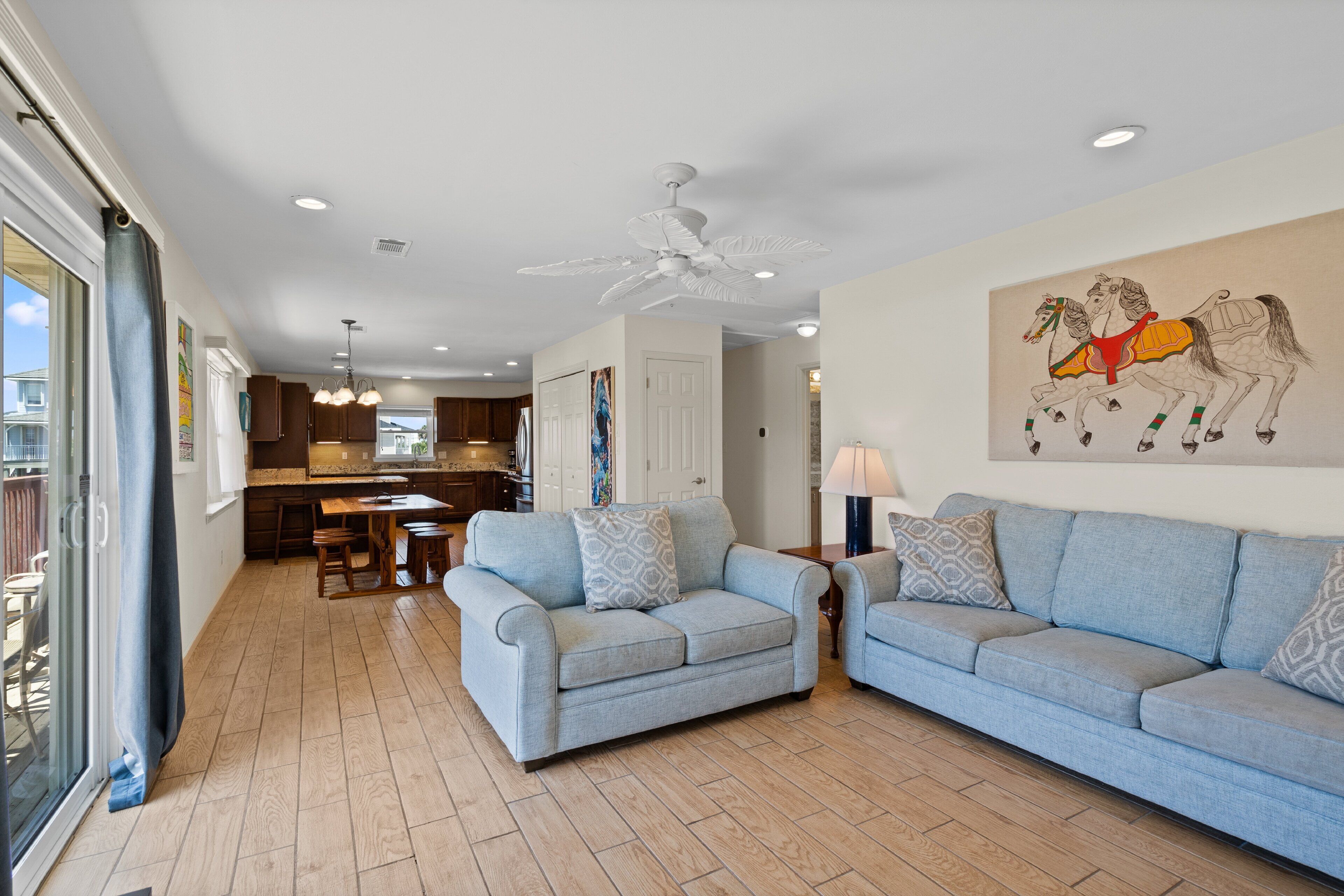 A spacious living room with two light blue sofas, a ceiling fan, and a kitchen in the background.