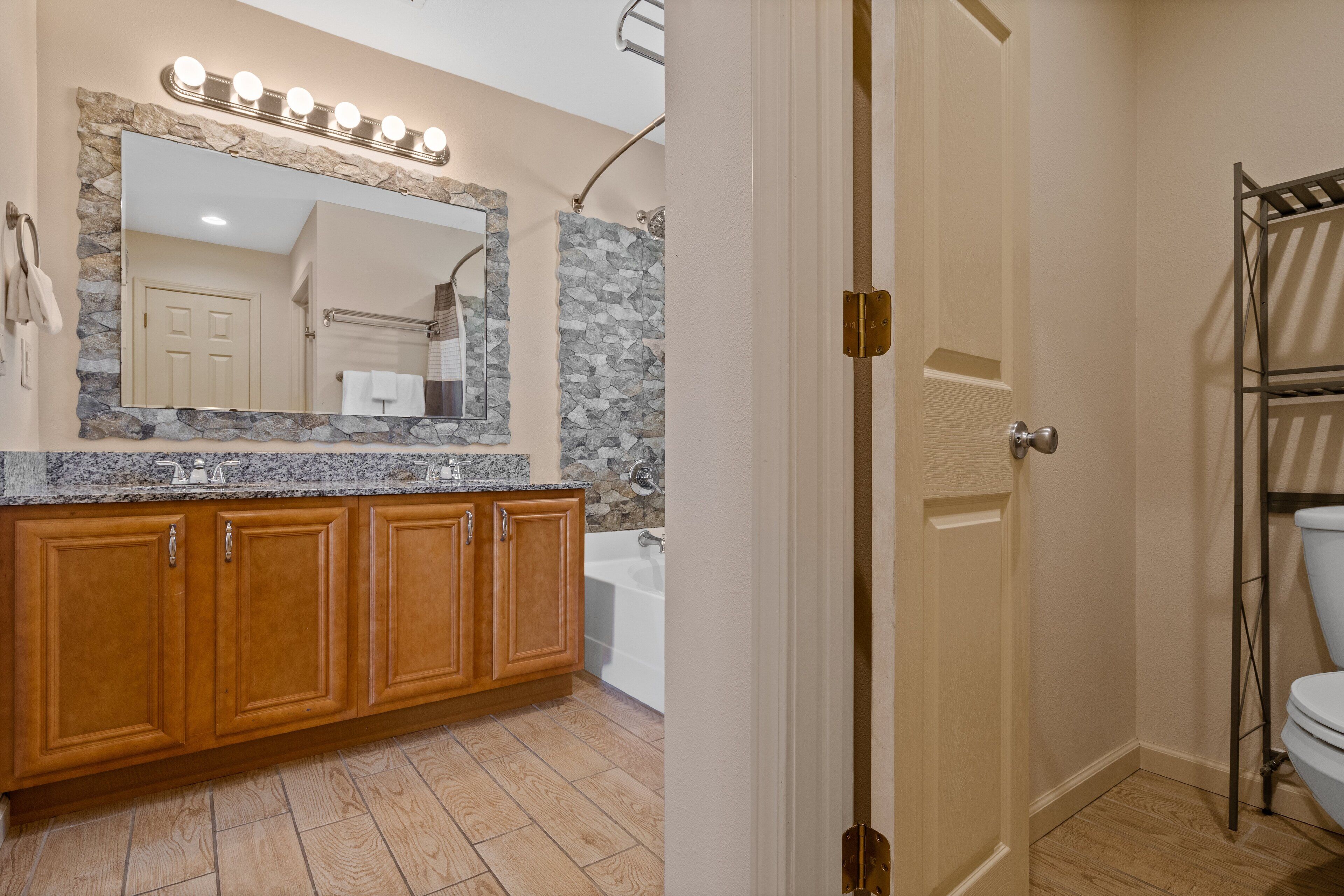 A bathroom with a wooden vanity, a large mirror with lights, a shower, and a toilet.