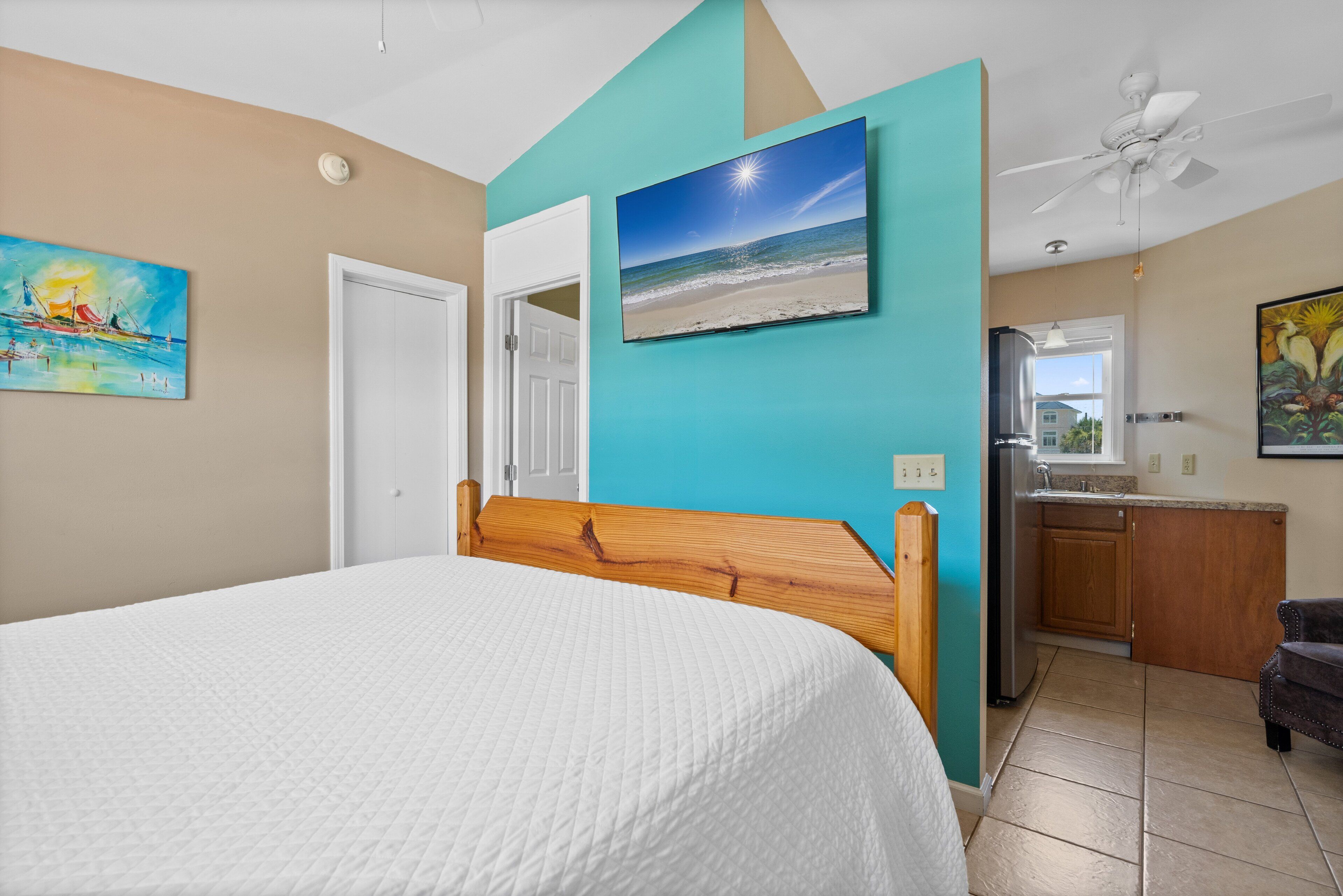 Une chambre à coucher avec un lit, un tableau représentant une scène de plage, une porte et une vue sur une cuisine et un balcon.