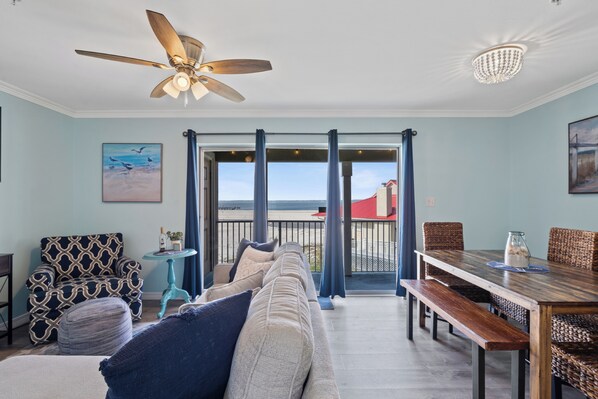 A living room with a sofa, a dining table with chairs, a ceiling fan, and a view through a glass door to the outside.