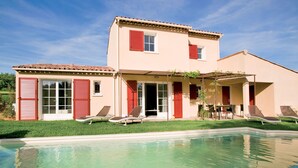 Une maison à deux étages avec une piscine, des volets rouges et une terrasse aménagée avec du mobilier d’extérieur.